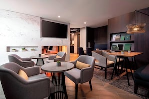 A modern lounge area with grey upholstered chairs, a round table, and a flat-screen TV mounted on the wall. - Vancouver Airport Marriott (Richmond)