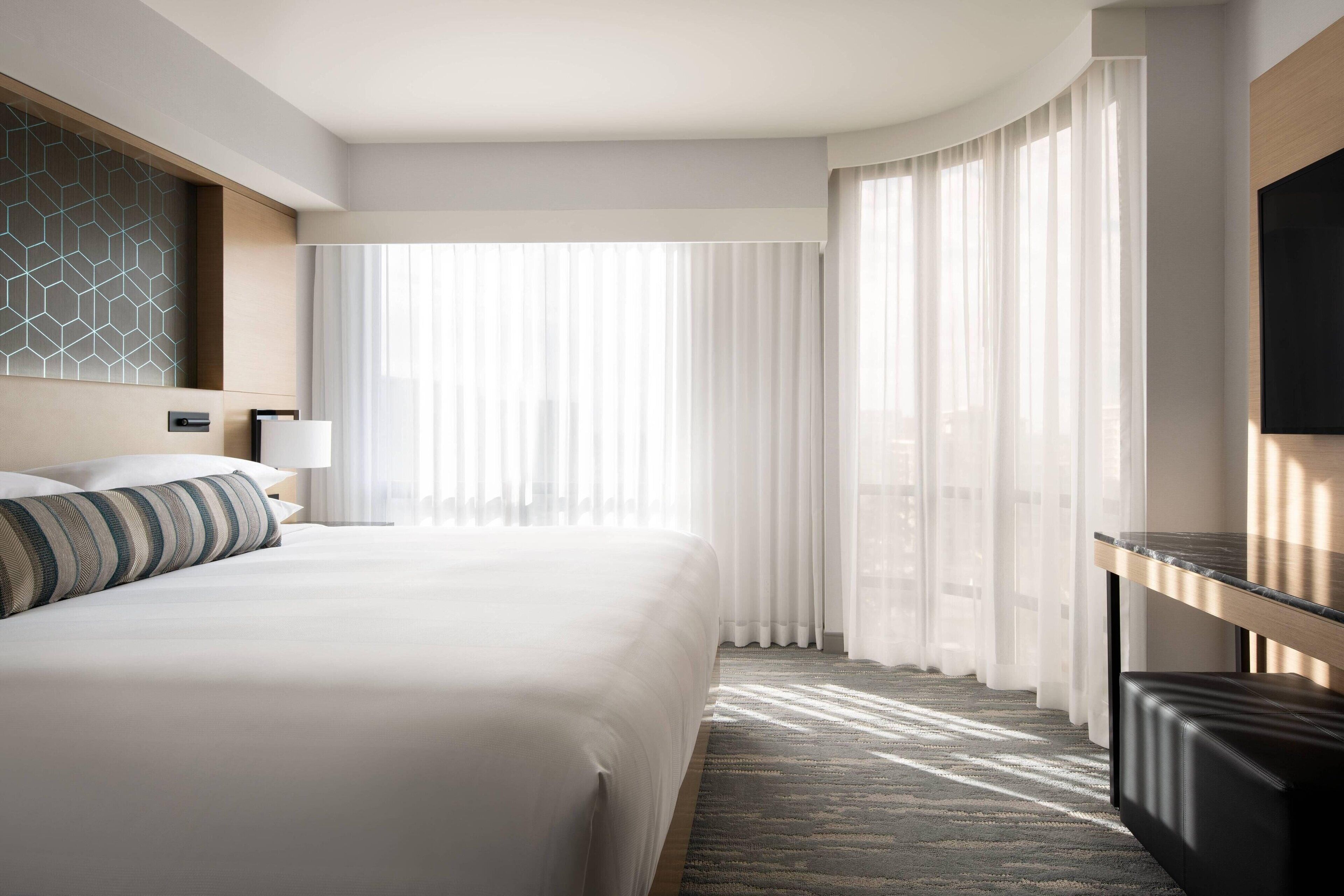 Habitación de hotel moderna con una cama grande, un escritorio y vistas a la ciudad a través de cortinas translúcidas.