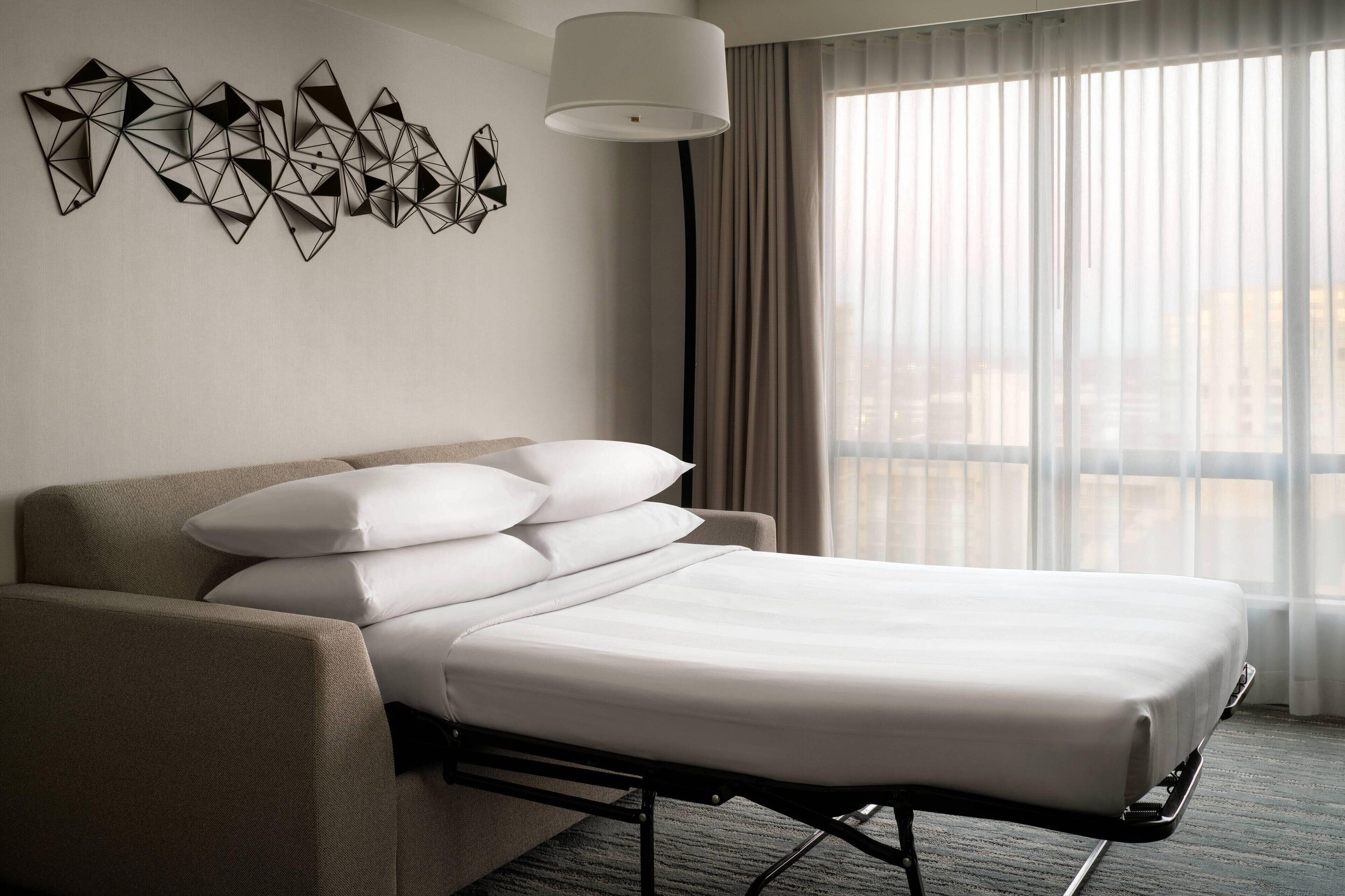 Quarto de hotel com cama, sofá, decoração geométrica na parede e janela com cortinas.