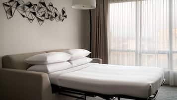Quarto de hotel com cama, sofá, decoração geométrica na parede e janela com cortinas.