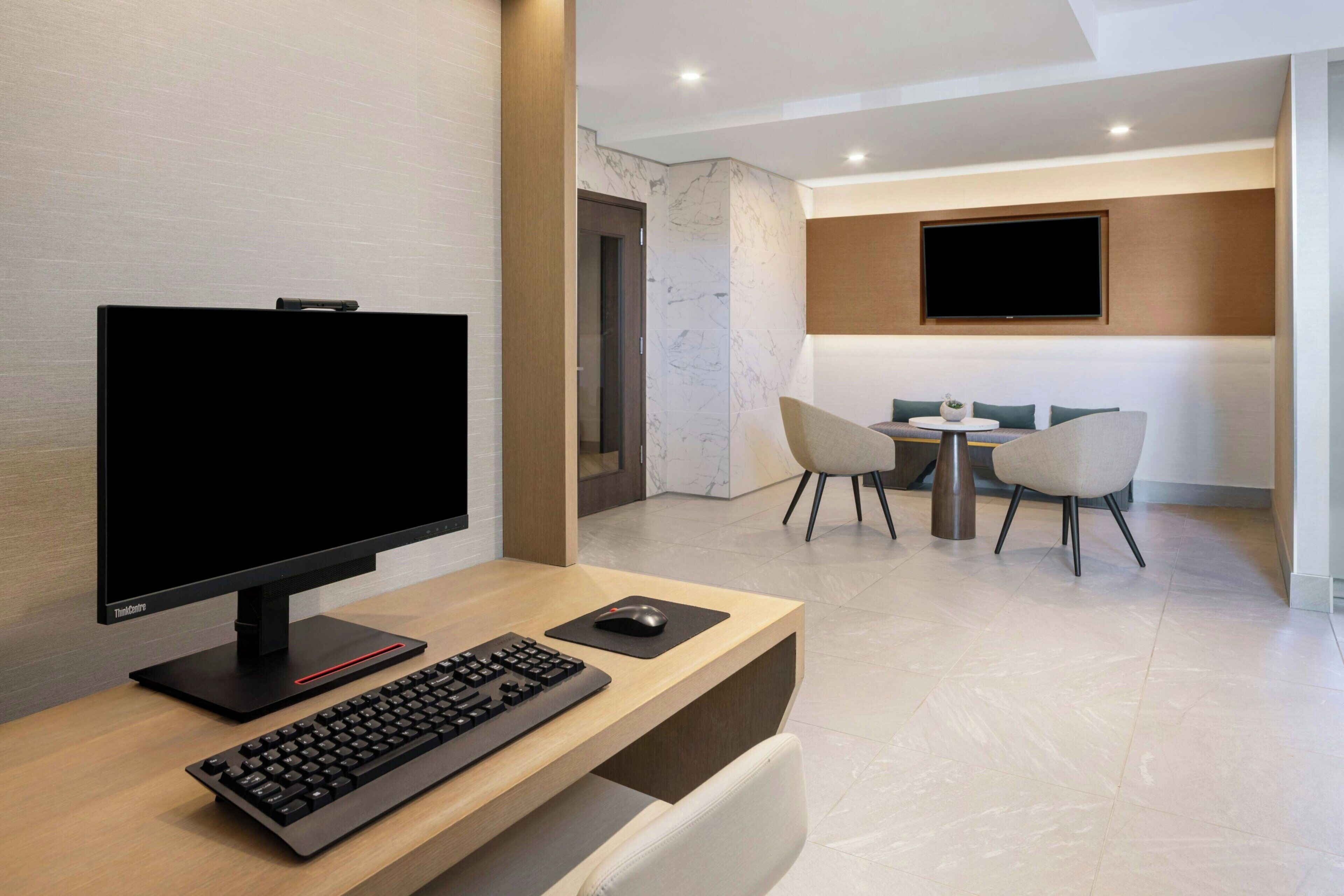 Um espaço de escritório moderno com mesa, computador, teclado e rato. Há uma televisão na parede, uma área de estar com cadeiras e uma porta ao fundo.