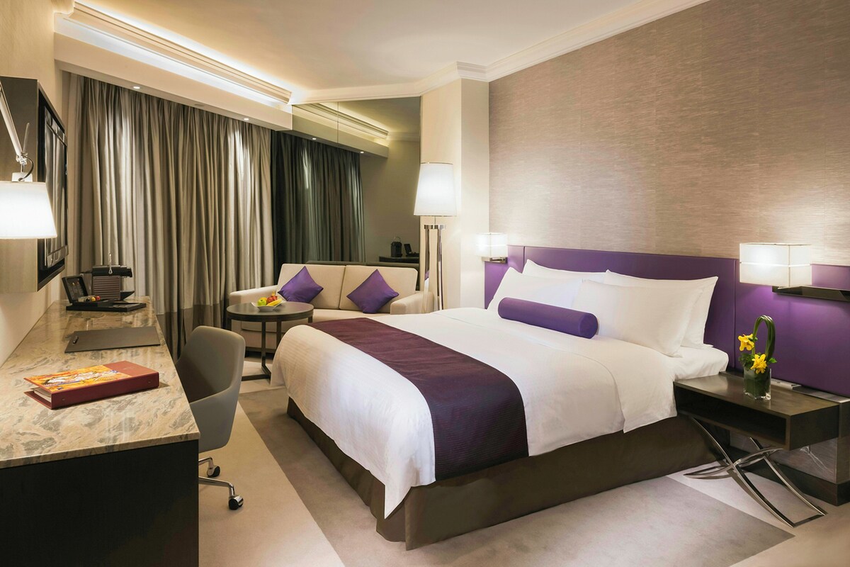 A hotel room with a bed, desk, chair, and a small table with a lamp.