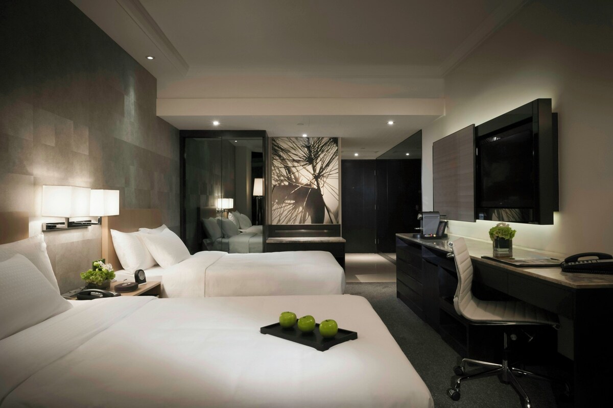 A hotel room with two beds, a desk, a chair, a TV, and a large window.