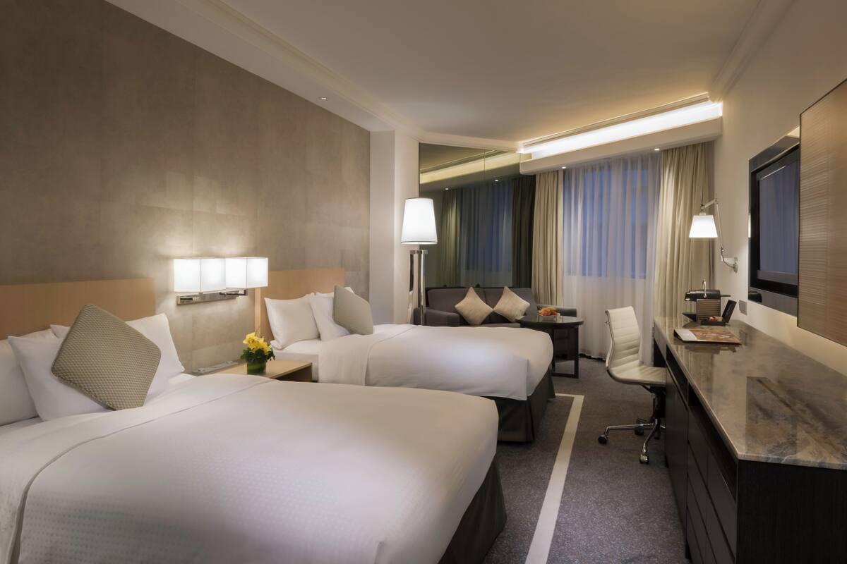 A hotel room with two beds, a desk, a chair, a TV, and a lamp.