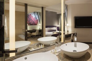 A modern hotel bathroom with two large vessel sinks, a large mirror, and a flat-screen TV mounted on the wall.
