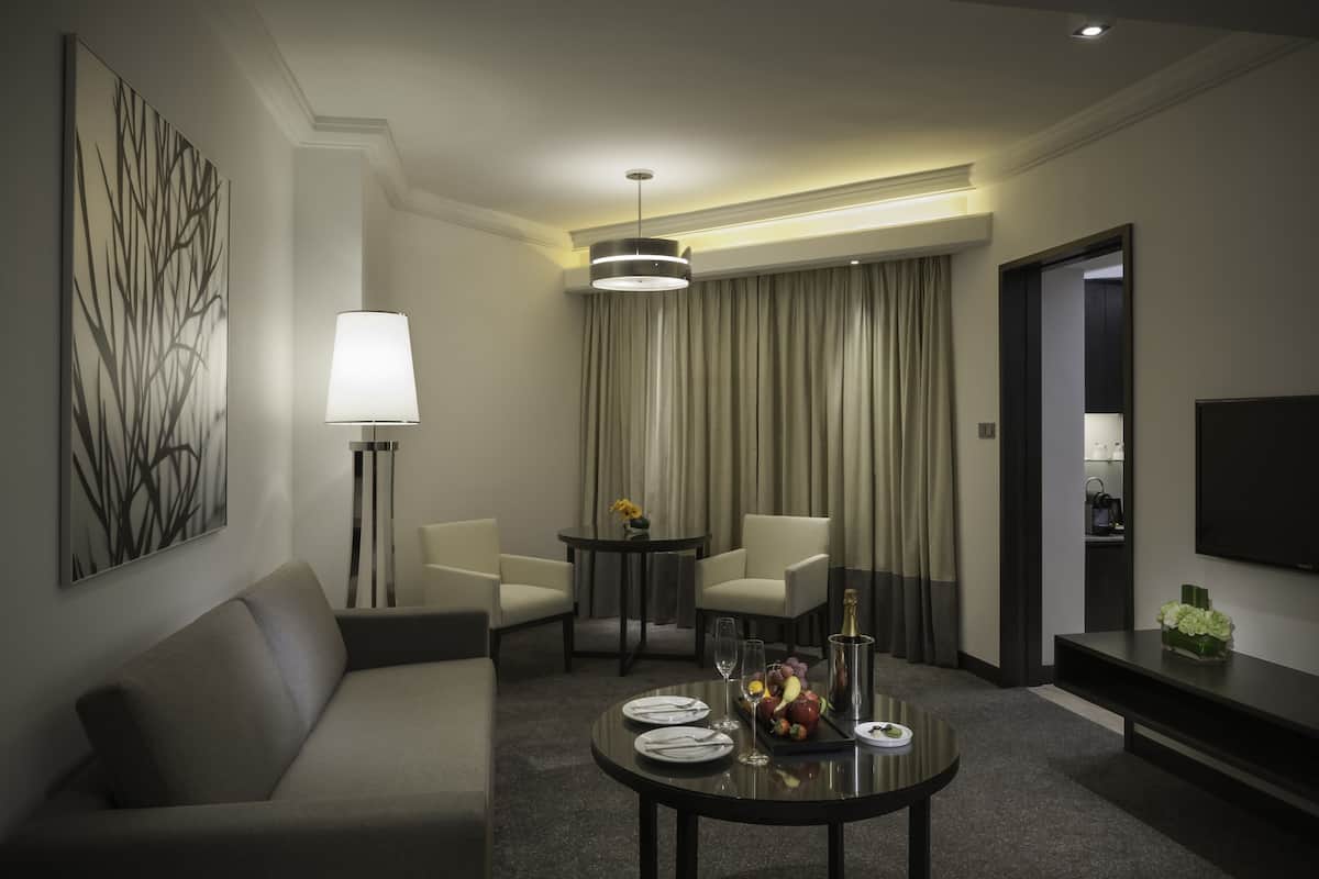 A modern hotel room with a sofa, a dining table set with plates, glasses, and a fruit bowl, a lamp, and a TV on the wall.