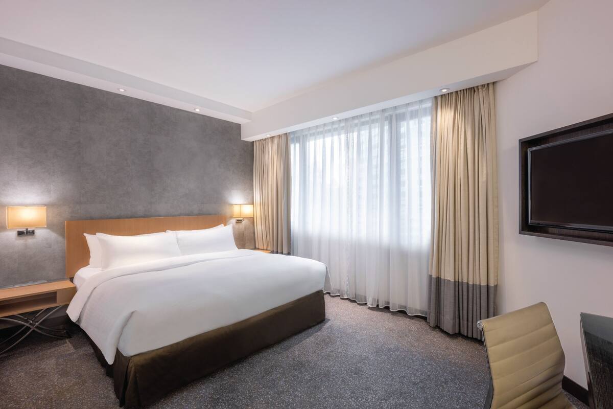 A hotel room with a large bed, two bedside lamps, a flat-screen TV, a chair, and a large window with curtains.