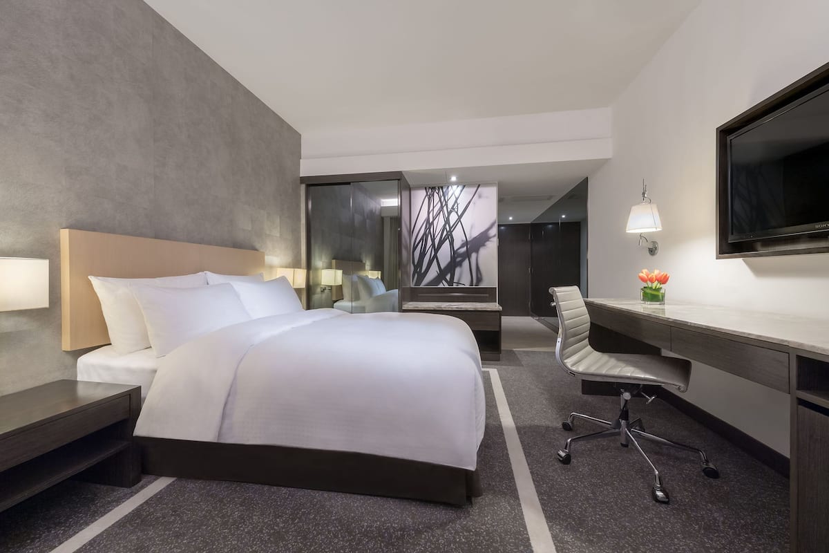A modern hotel room with a large bed, a desk, a chair, a TV, and a wall-mounted lamp.