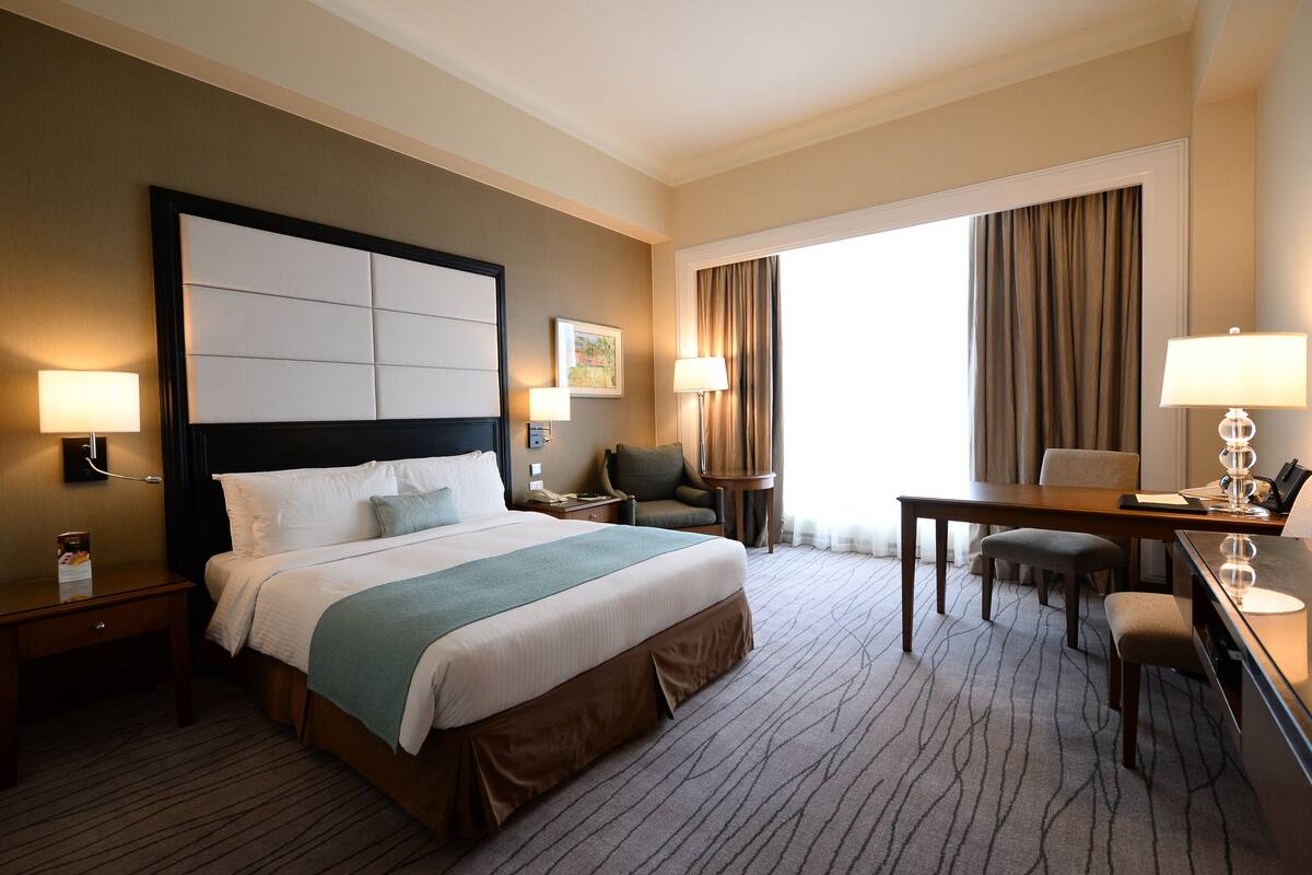 Quarto de hotel com cama grande, escrivaninha, duas cadeiras, mesa pequena e janela com cortinas.