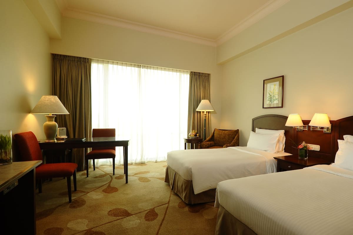 Quarto de hotel com duas camas, uma escrivaninha, uma cadeira e um abajur.