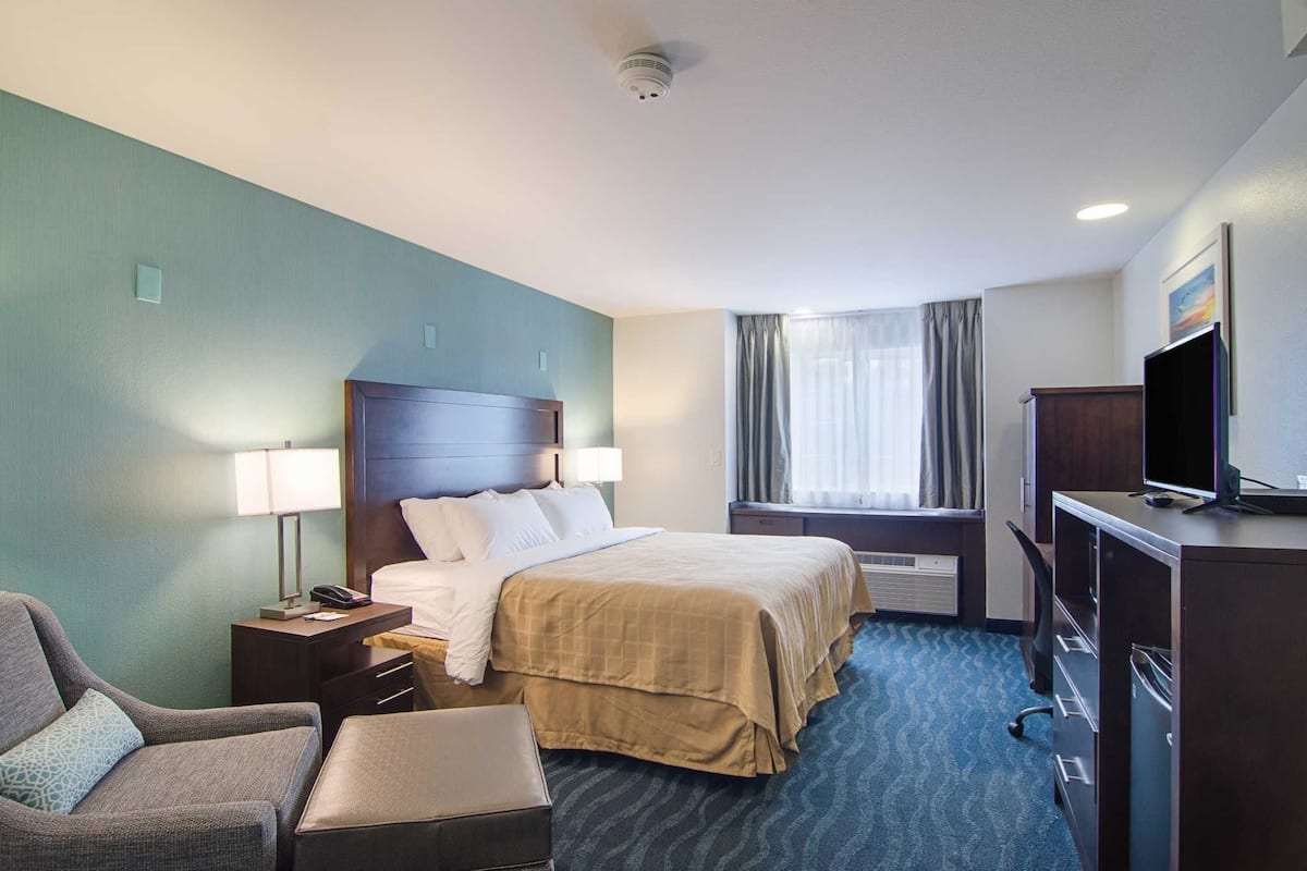 A hotel room with a bed, a desk with a computer, a chair, a TV, and a window with curtains.