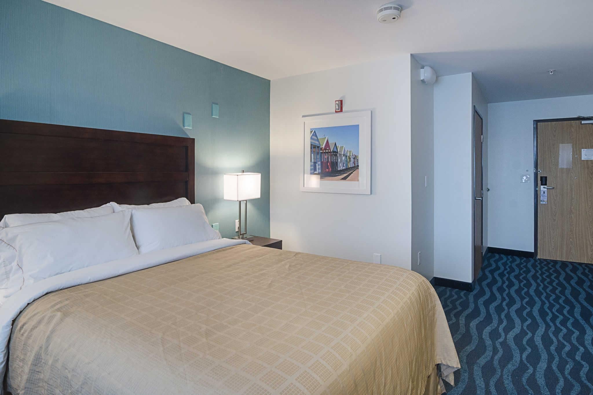 A hotel room with a large bed, a nightstand with a lamp, a framed picture on the wall, and a door to the right.