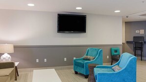 A modern hotel room with a flat-screen TV mounted on the wall, two teal armchairs, a white coffee table, and a small side table with a lamp.