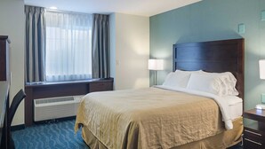 A hotel room with a bed, a desk, a chair, a window with curtains, a wall-mounted lamp, and a heater.