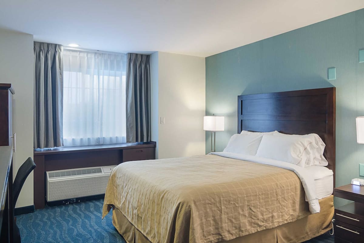 A hotel room with a bed, a desk, a chair, a window with curtains, a wall-mounted lamp, and a heater.