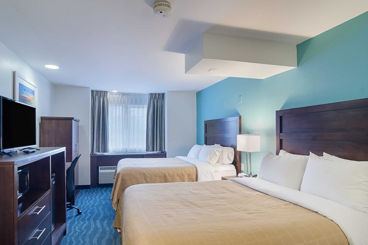 A hotel room with two beds, a desk with a television, a window with curtains, and a ceiling-mounted security camera.