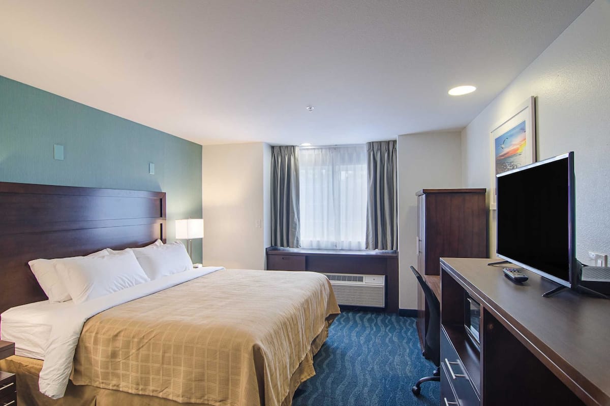 A hotel room with a bed, a desk with a TV, a window with curtains, and a painting on the wall.