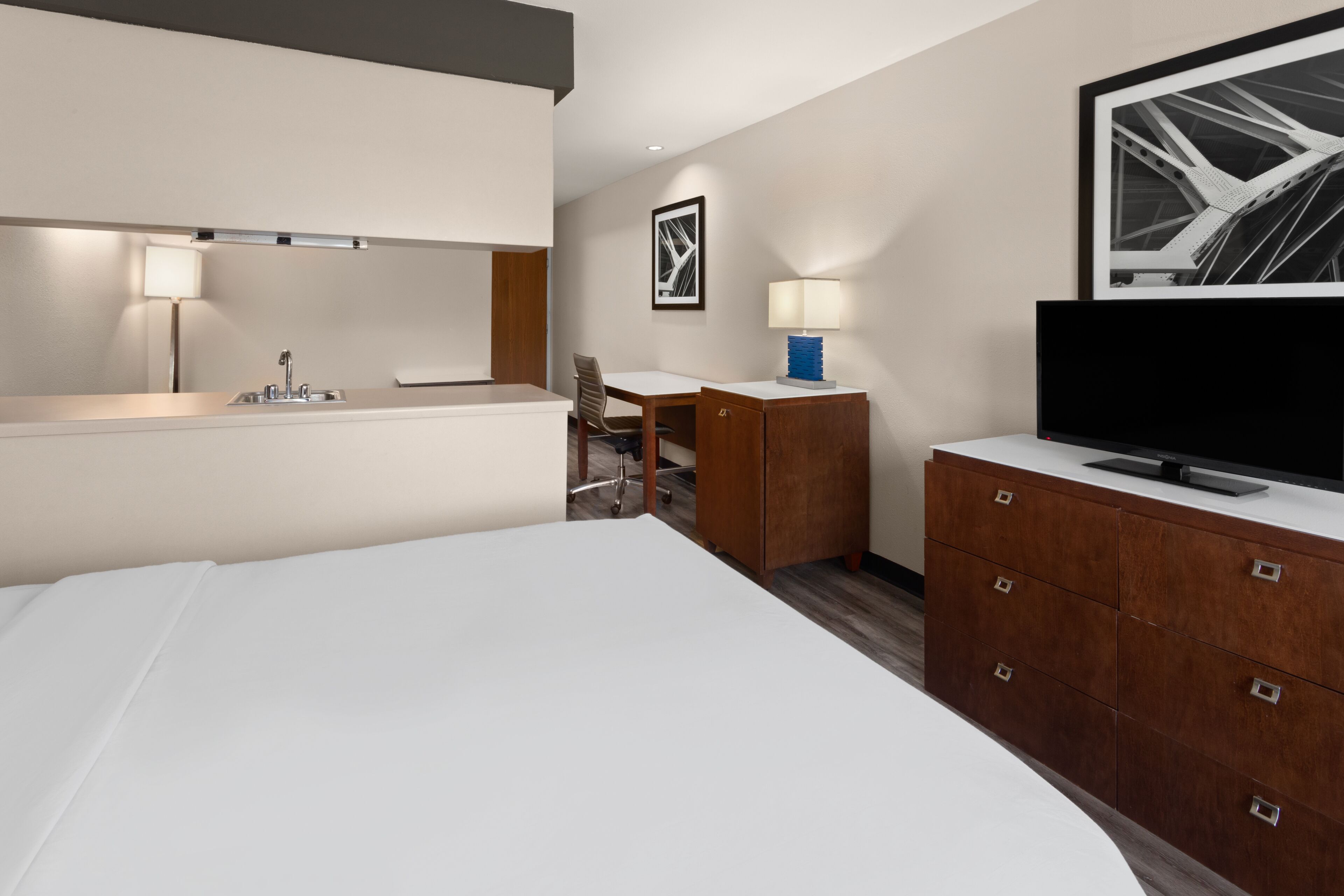 A hotel room with a bed, a desk, a chair, a TV, a lamp, and a framed picture on the wall.