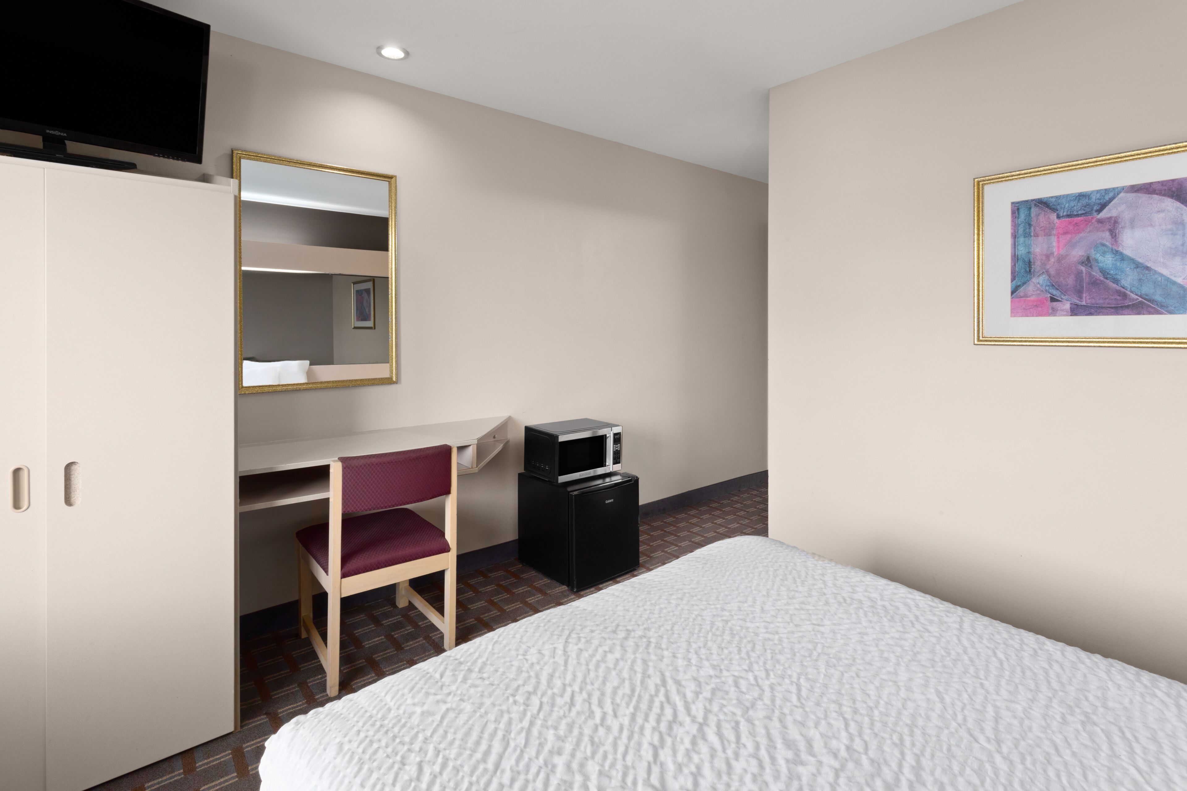 A hotel room with a bed, a desk with a chair, a television, a microwave, and a framed picture on the wall.