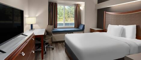 A hotel room with a large bed, a desk with a TV, a window with curtains, and a view of trees.