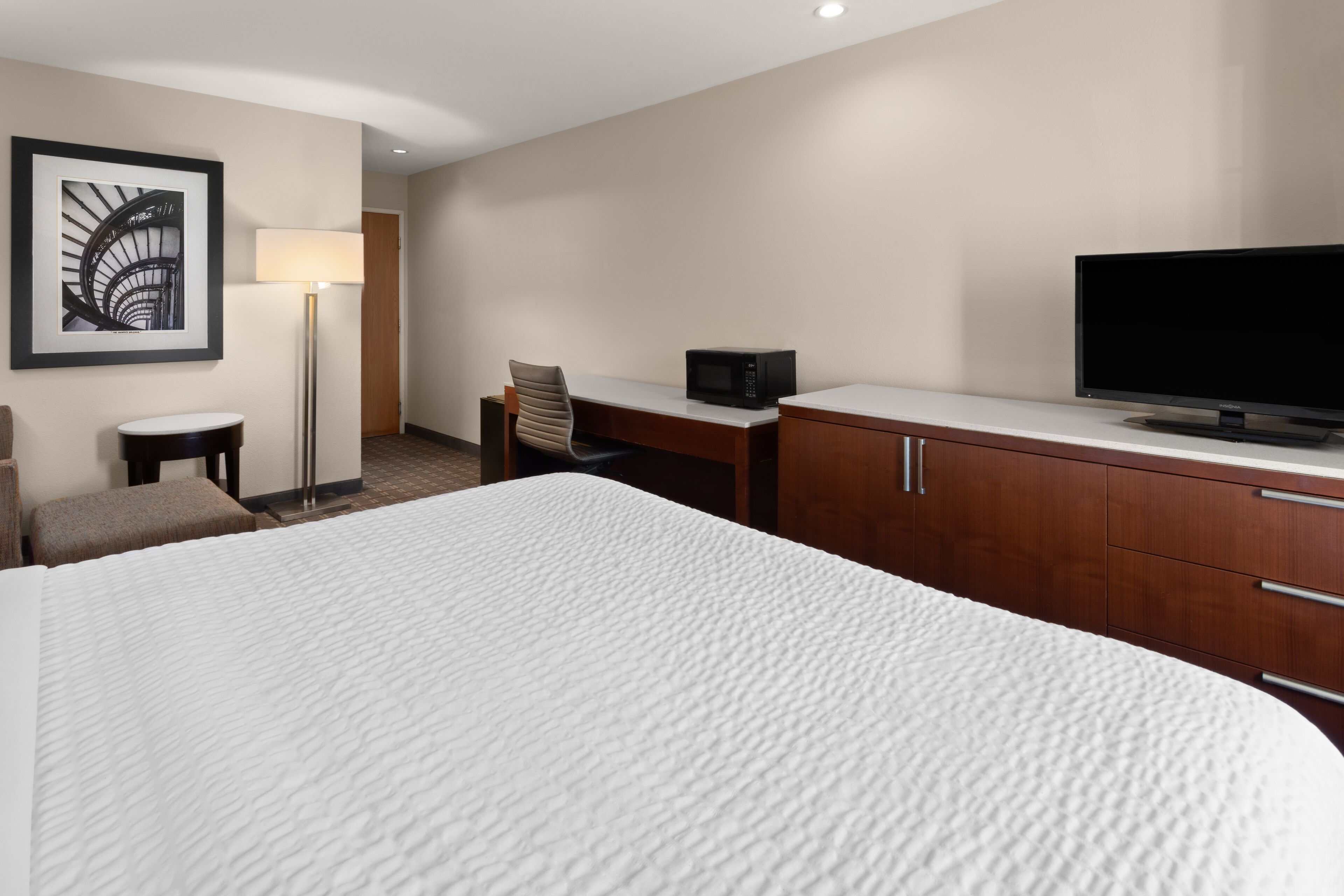 A hotel room with a large bed, a desk with a chair, a television, a lamp, and a framed picture on the wall.