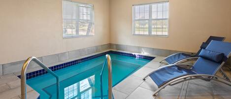 Una piscina con borde de azulejos azules, dos ventanas con persianas y sillones de descanso con cojines azules.