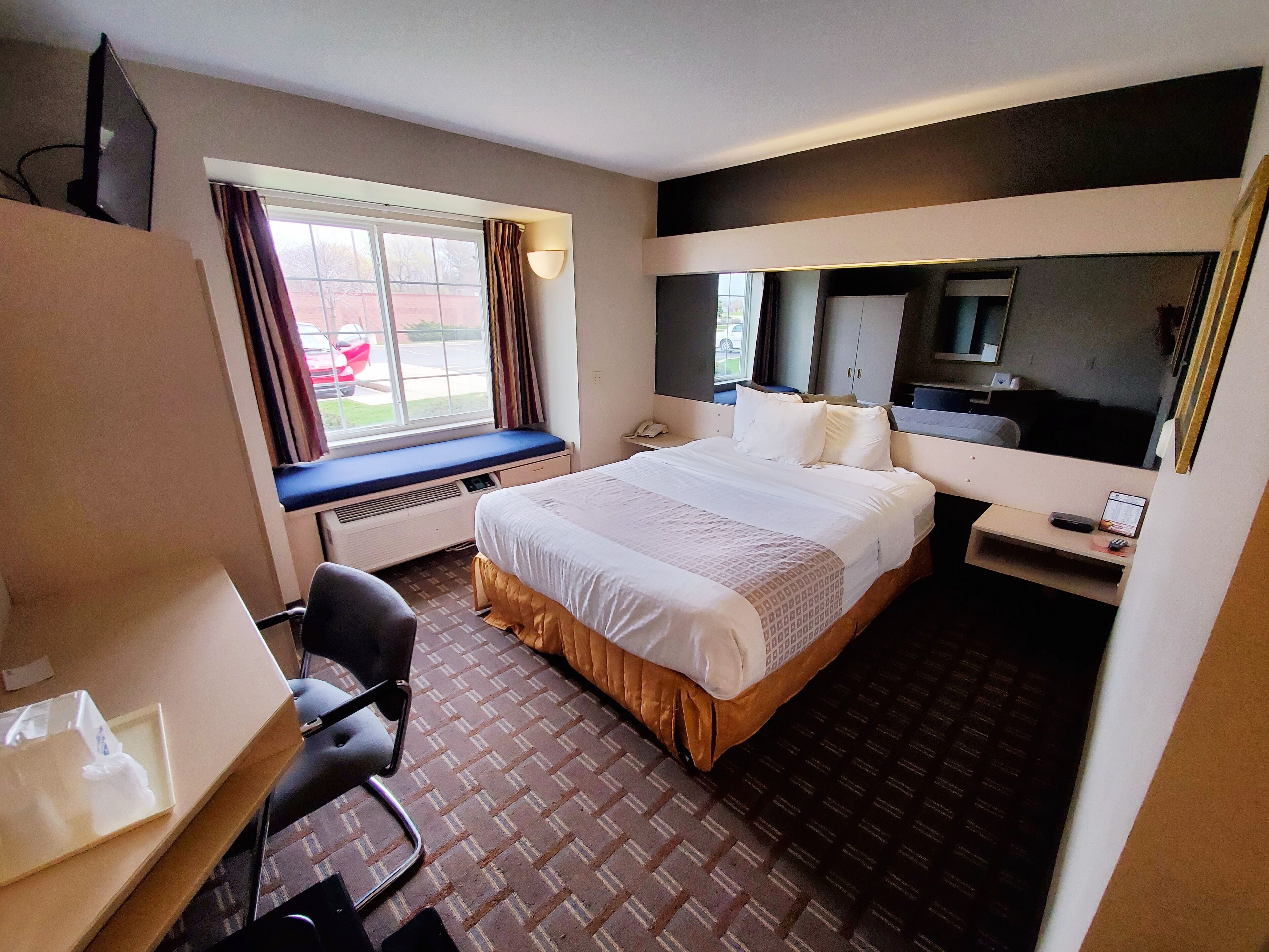 A hotel room with a large bed, a desk with a chair, a window with curtains, a bedside table with a phone, and a mirror.