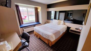 A hotel room with a large bed, a desk with a chair, a window with curtains, a bedside table with a phone, and a mirror.