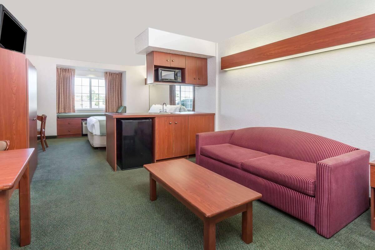Ein kompaktes Hotelzimmer mit Küchenzeile, einer Couch, einem Holztisch und einem Bett mit Blick aus dem Fenster.