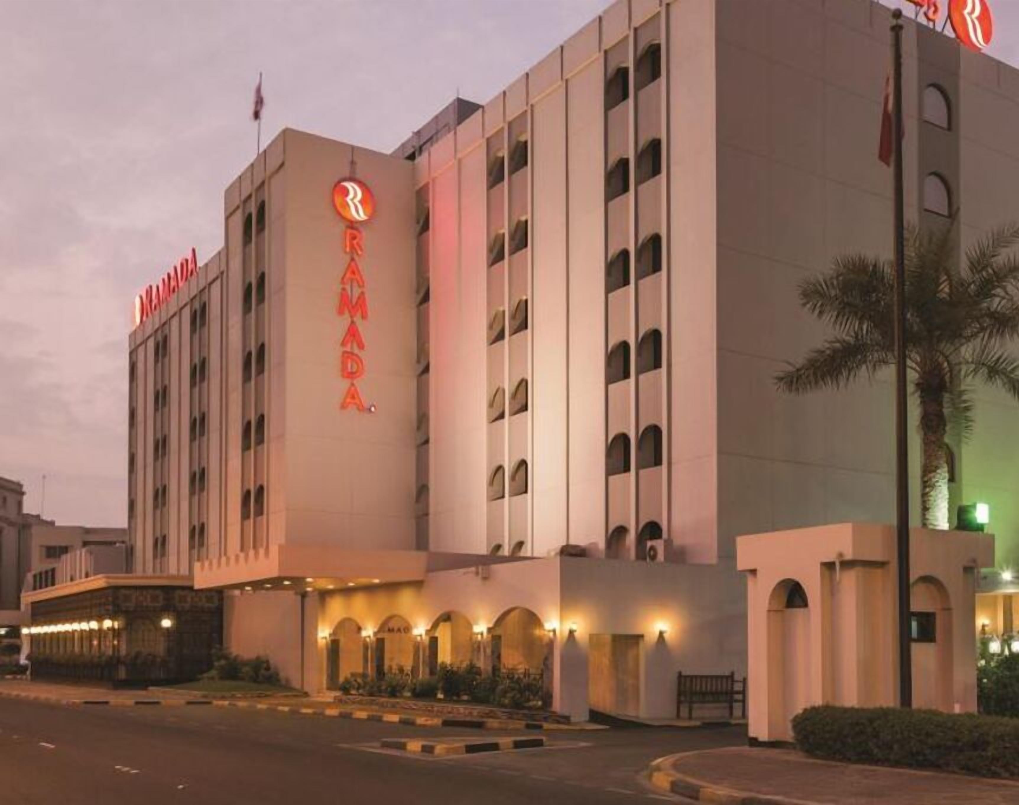 A hotel with a prominent 'RAMADA' sign, multiple floors, and a well-lit entrance with a palm tree nearby.