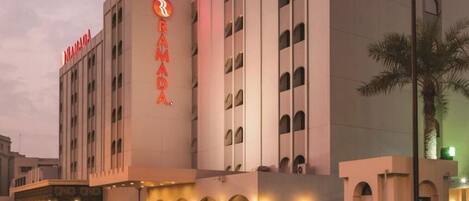 A hotel with a prominent 'RAMADA' sign, multiple floors, and a well-lit entrance with a palm tree nearby.