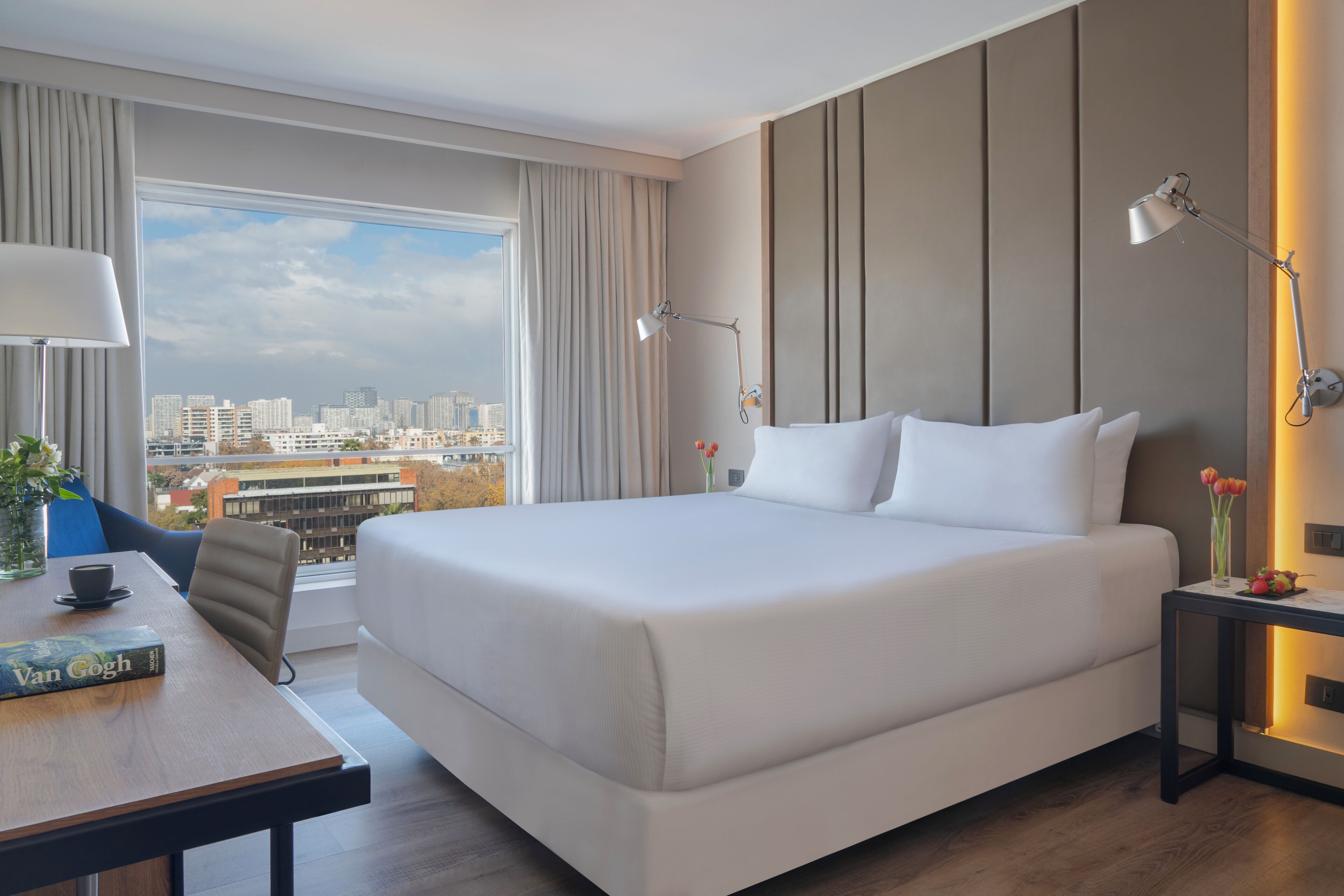 A hotel room with a large bed, a desk with a chair, a lamp, and a view of the city.
