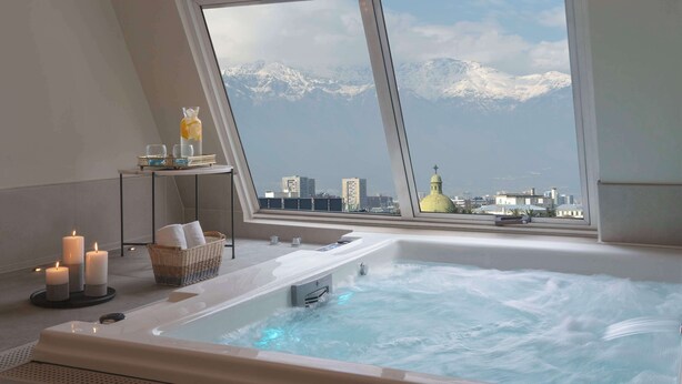 Une salle de spa dotée d’une grande baignoire, de bougies, d’un panier et offrant une vue sur des montagnes enneigées et un paysage urbain, le tout visible à travers une grande fenêtre.