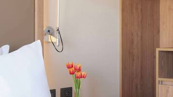 Una cama bien tendida con sábanas blancas, una lámpara de noche, una mesita con un jarrón de tulipanes y un armario de madera con perchas.