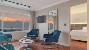 Une chambre d’hôtel avec un grand lit, deux fauteuils bleus, une table basse ronde, un téléviseur à écran plat et une grande fenêtre donnant sur la montagne.