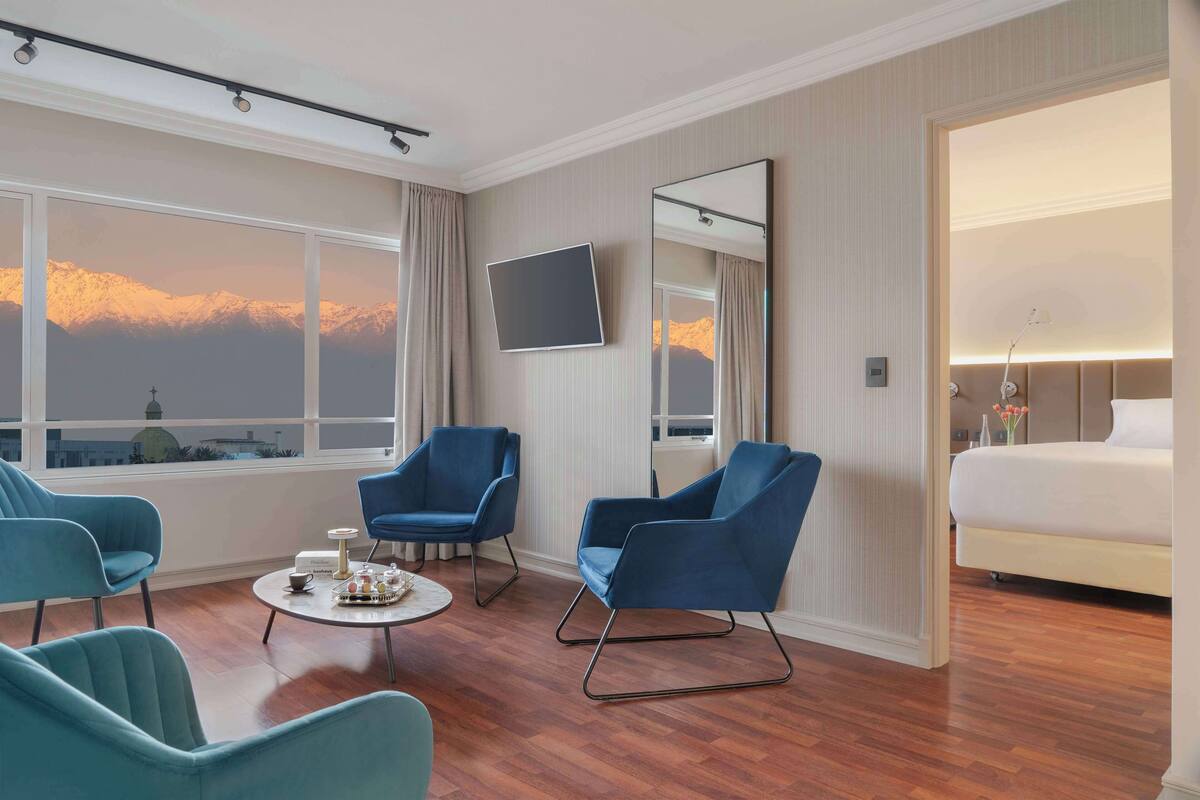 Une chambre d’hôtel avec un grand lit, deux fauteuils bleus, une table basse ronde, un téléviseur à écran plat et une grande fenêtre donnant sur la montagne.