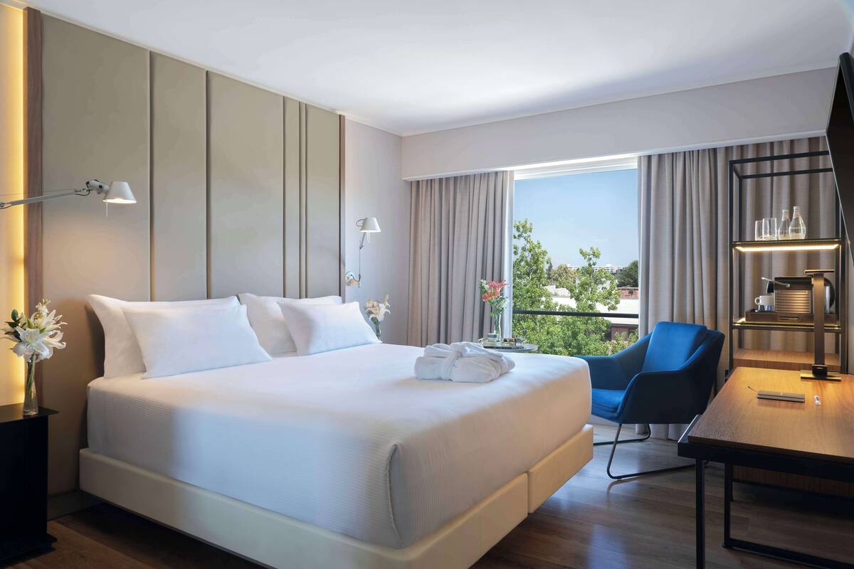 Une chambre d’hôtel moderne avec un grand lit, un bureau et une chaise. On y trouve une fenêtre avec des rideaux, un vase avec des fleurs et une vue sur la verdure à l’extérieur.