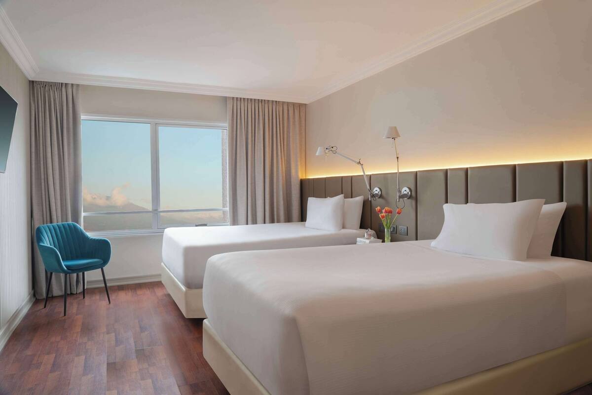Une chambre d’hôtel avec deux lits, une chaise, une fenêtre avec des rideaux et une lampe de chevet.