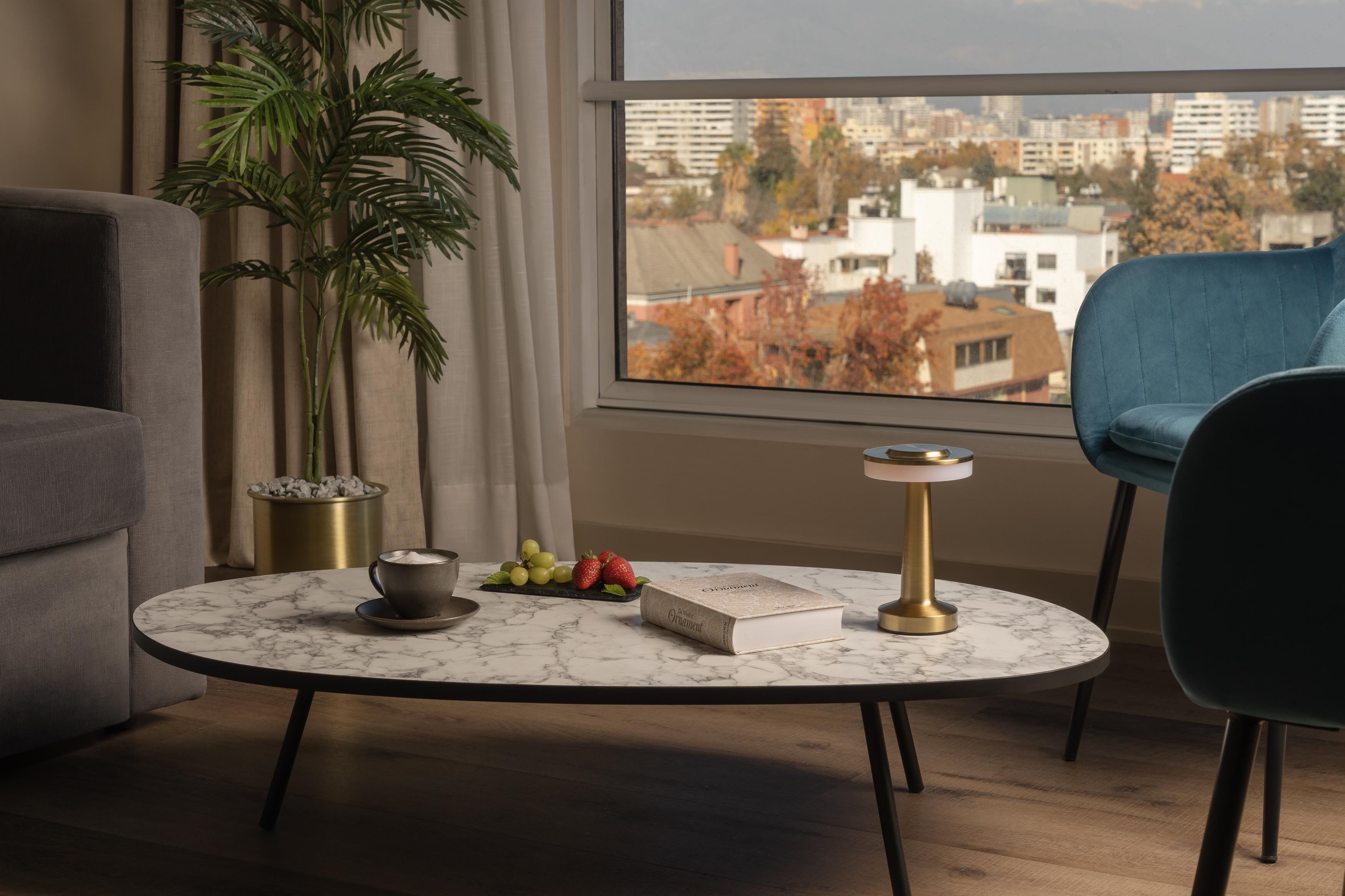 Un salon moderne avec une table basse ronde en marbre, un livre, une théière et un panier de fruits. Une grande fenêtre offre une vue sur le paysage urbain.