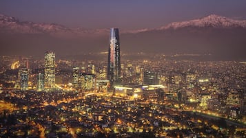 Un paisaje urbano nocturno con un rascacielos destacado, una zona urbana densa y montañas nevadas al fondo.