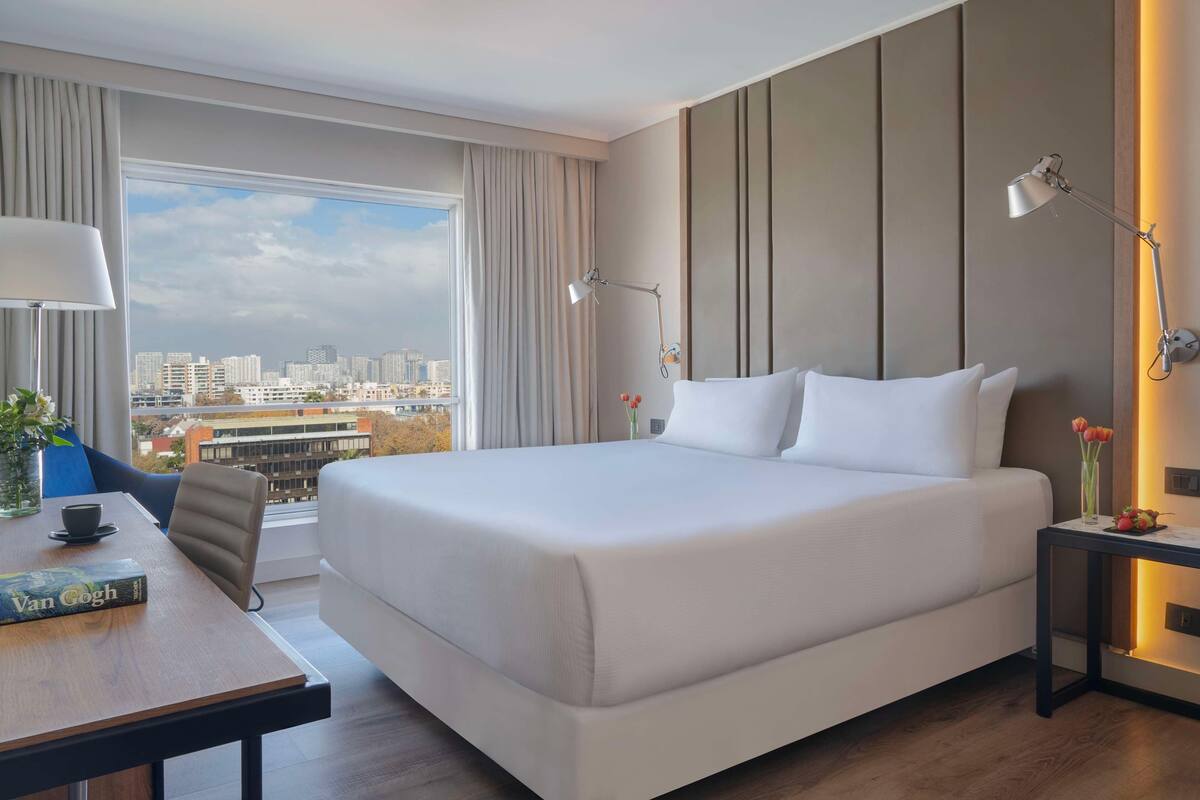 Une chambre d’hôtel avec un grand lit, un bureau avec un livre, une lampe et une vue sur le paysage urbain.