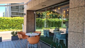 Un área de estar al aire libre con mesas y sillas redondas, una pared de vidrio con vegetación y una fachada de edificio moderna.