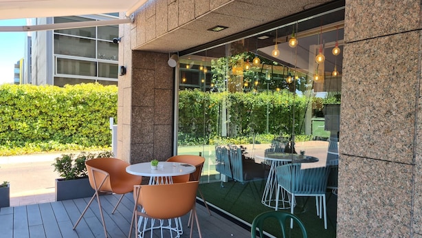 Un espace extérieur aménagé avec des tables et des chaises rondes, un mur de verre agrémenté de verdure et une façade de bâtiment moderne.