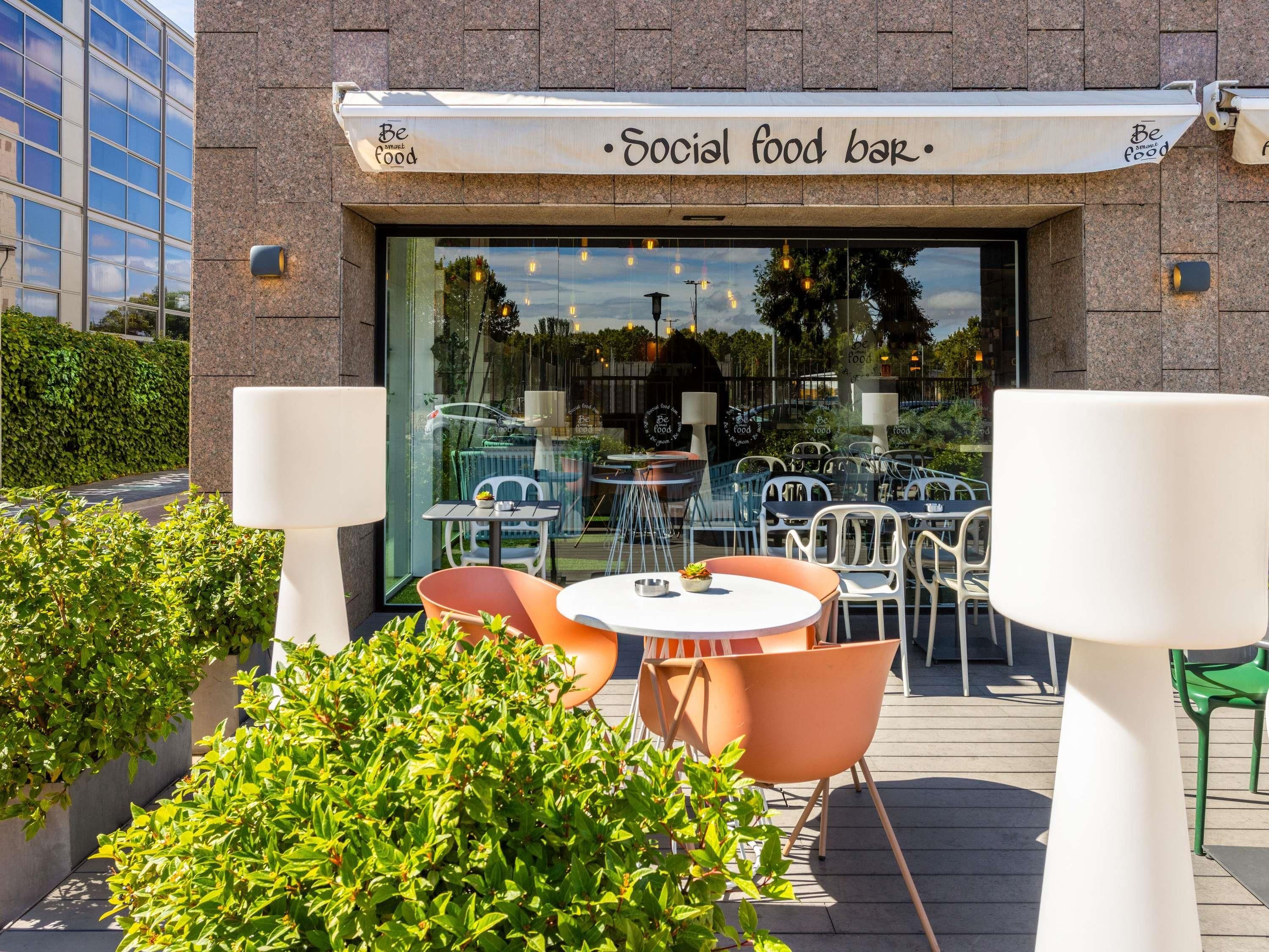 An outdoor seating area with tables and chairs, a social food bar, and a glass facade reflecting the surroundings.