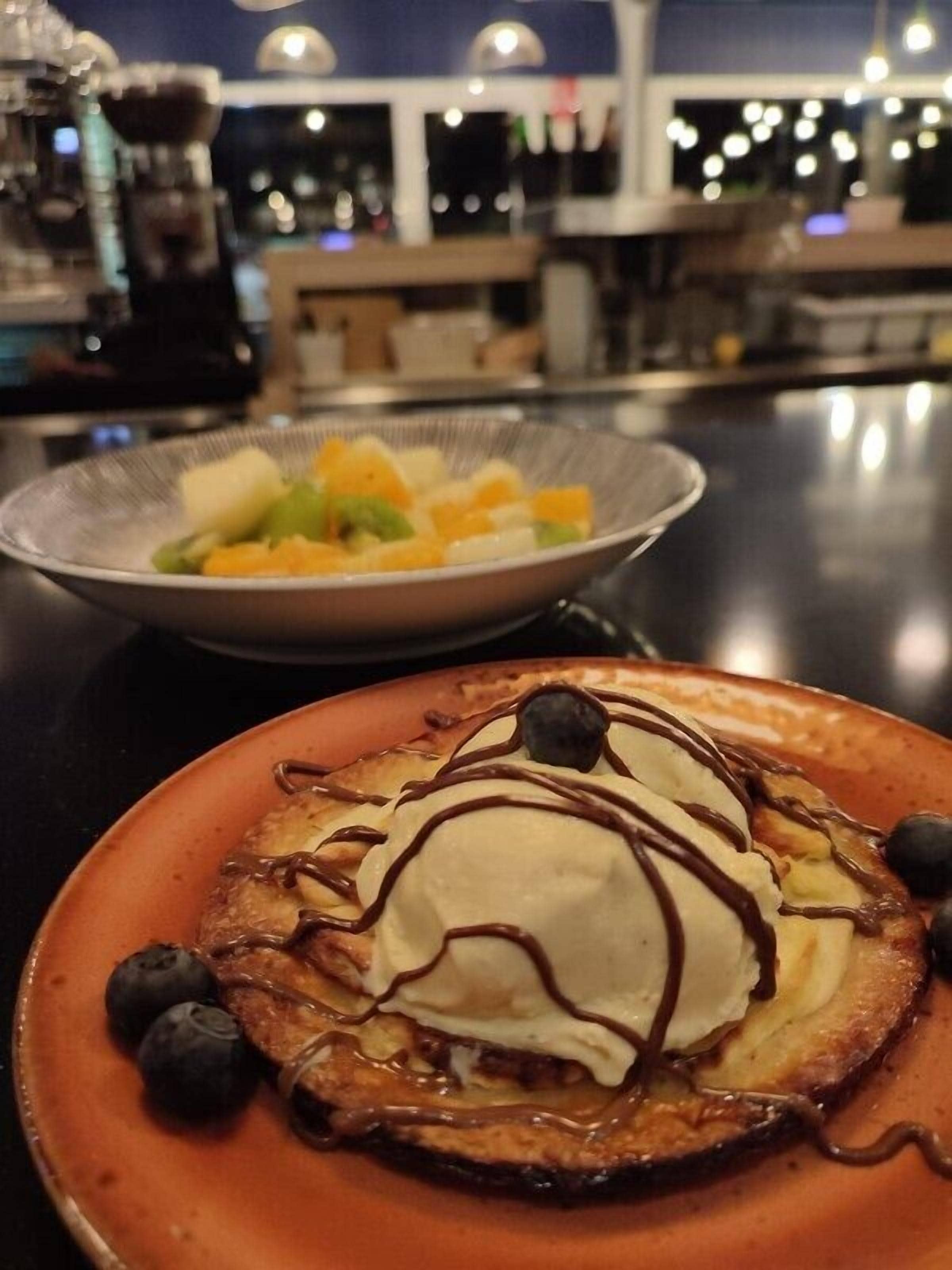 Un plato de postre con tortitas, helado y salsa de chocolate, y un bol de fruta al fondo.