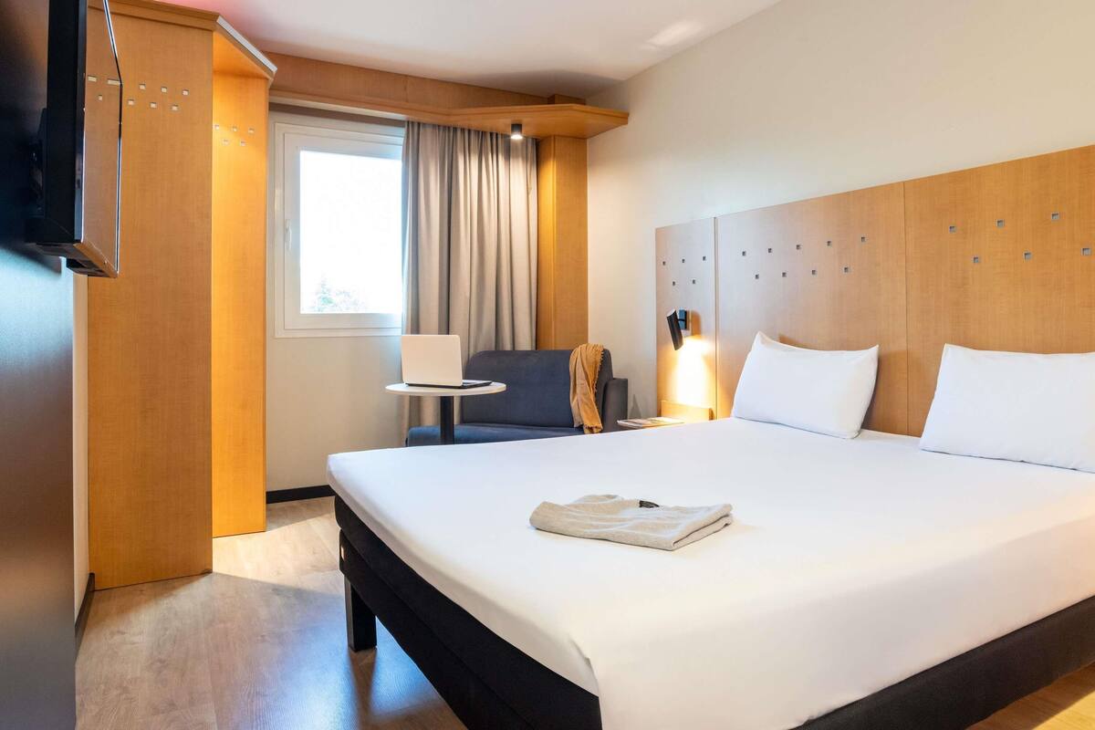 Una habitación de hotel con una cama grande, un escritorio con una computadora portátil, una silla y un televisor montado en la pared.