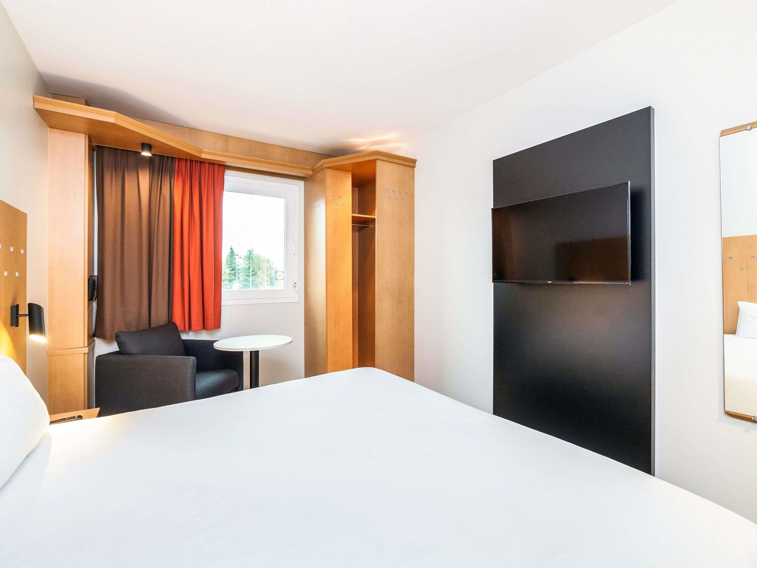 A hotel room with a bed, a TV mounted on the wall, a small table, and a seating area with a chair.