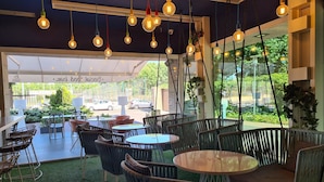 Un café al aire libre con mesas y sillas redondas, lámparas colgantes y vistas a la calle.