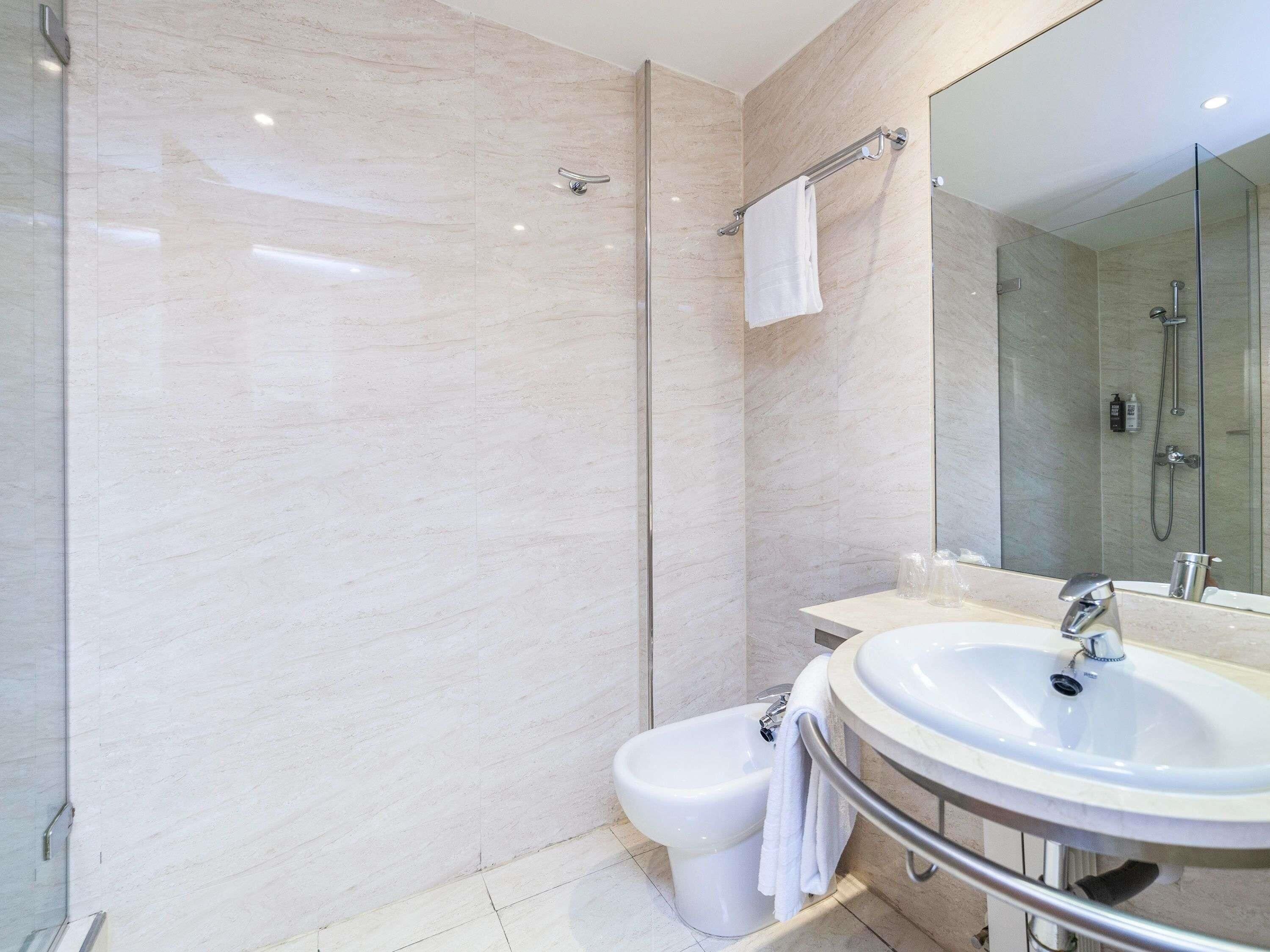 A modern bathroom with a marble wall, a glass shower enclosure, a white sink, and a bidet.