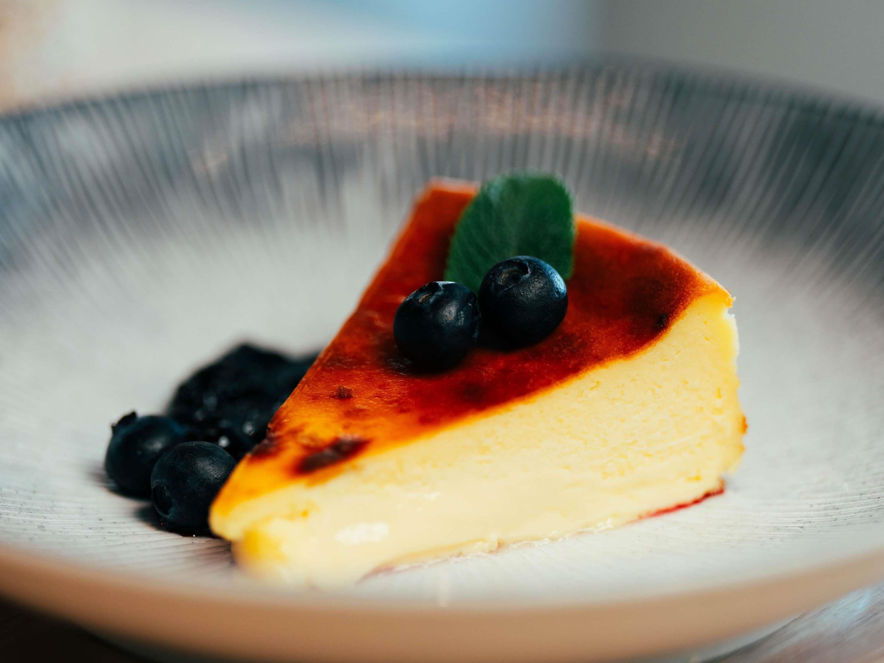 A slice of cheesecake with a caramelized top, blueberries, and a mint leaf on a plate.